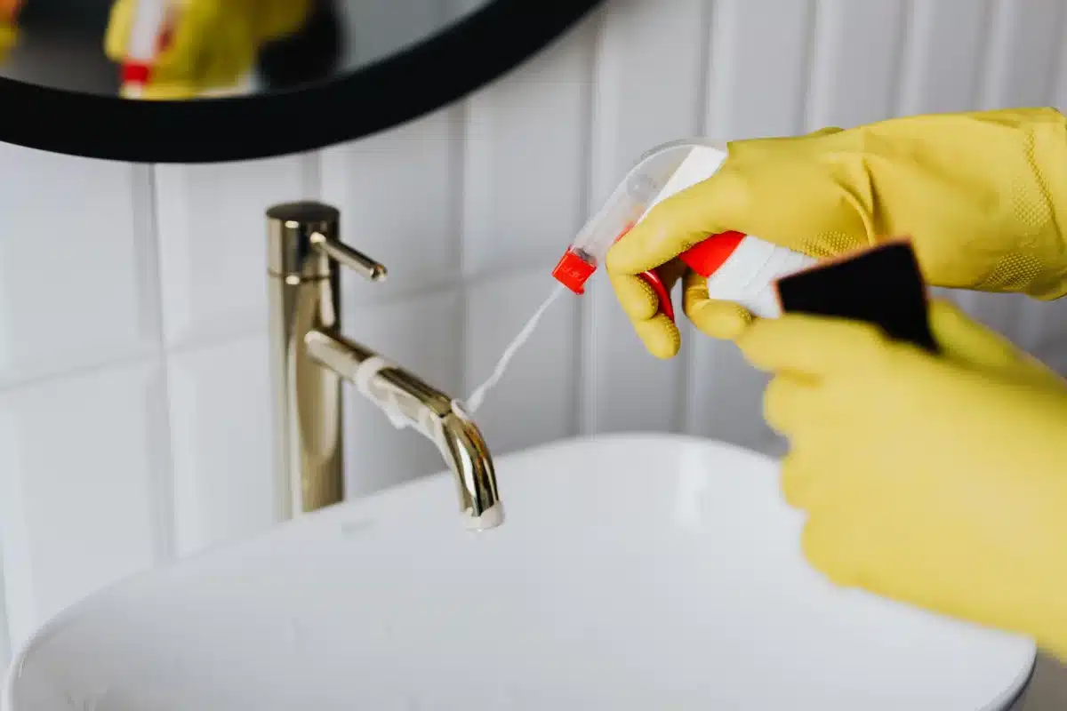 Hand in rubber gloves spraying cleaner on a chrome bathroom faucet - How to Deep Clean a Bathroom: A Room-by-Room Checklist